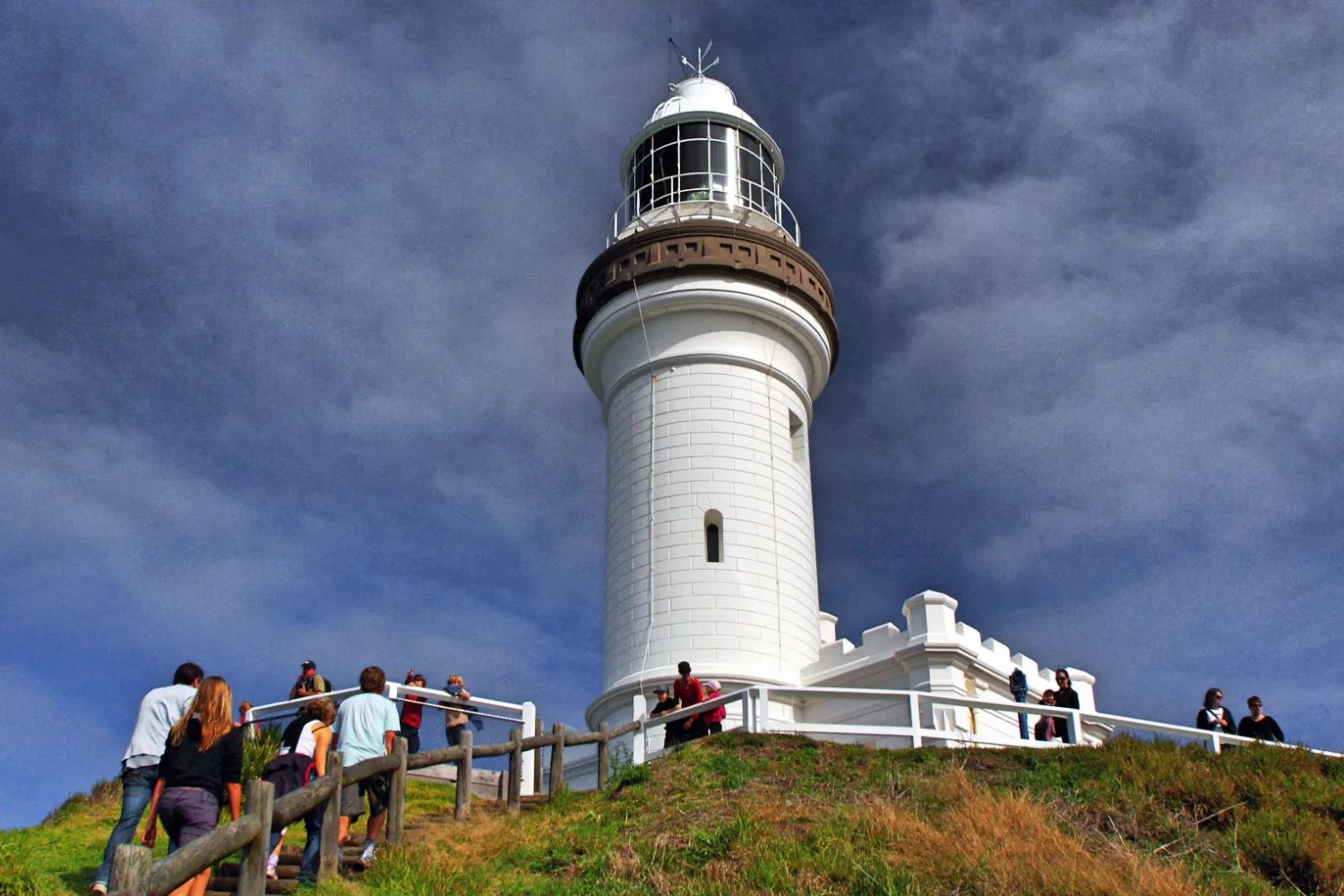 The white Cape Byron lighthouse on the headland, with walkers at the rail on the path below