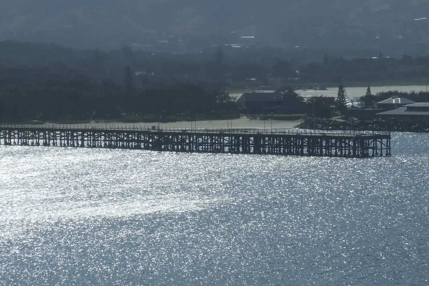 The long timber jetty at Coffs Harbour on its piles over glittering water under a grey sky, with the town and wooded hills behind
