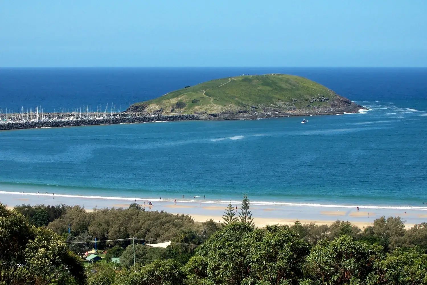 Muttonbird Island seen from the shore: a low green island at the end of a rock breakwall, with yachts in the marina and deep blue sea behind