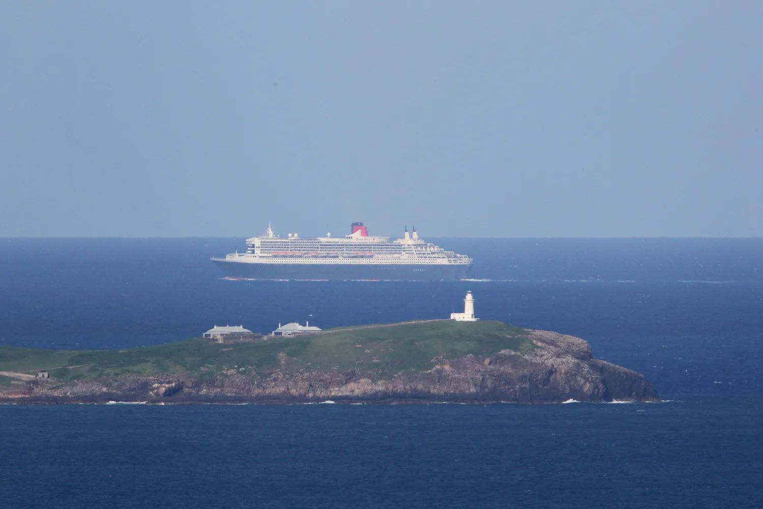 South Solitary Island and its white lighthouse on a dark blue sea, with a large cruise ship passing on the horizon beyond