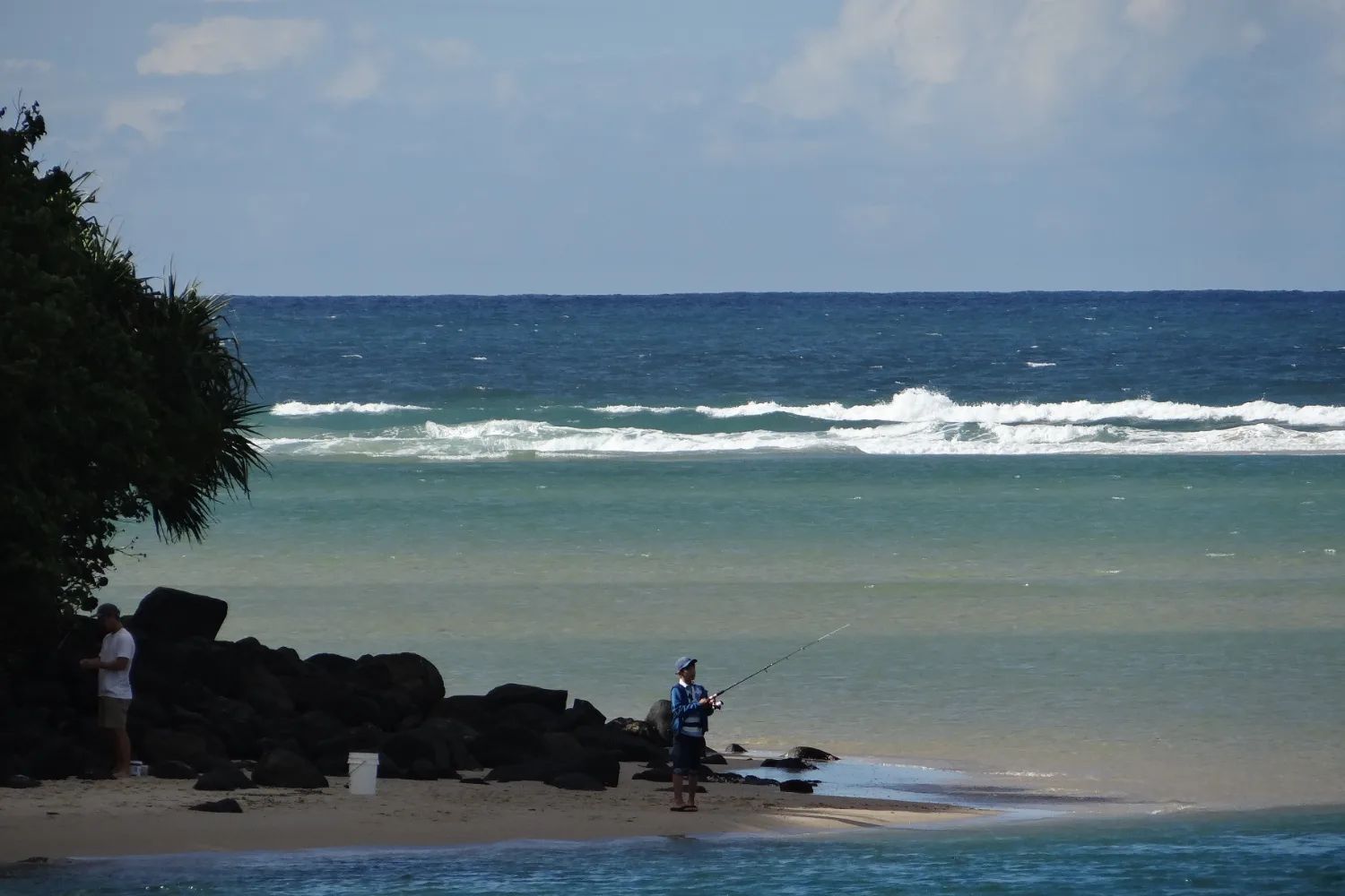 A fisherman on the black rocks at the foot of the Burleigh headland, with the rainforest edge on the left and surf beyond