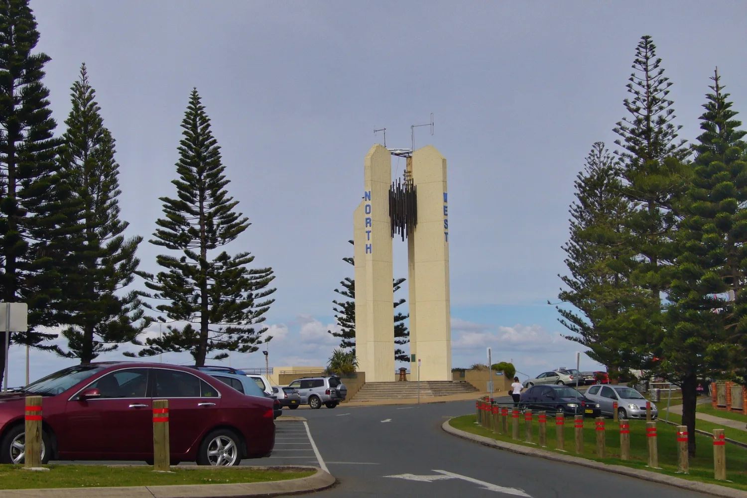 The tall white Captain Cook Memorial lighthouse at Point Danger, framed by Norfolk Island pines, with a car park in front