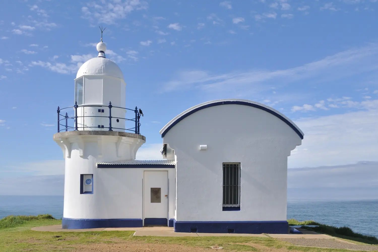 The small white tower of Tacking Point Lighthouse and its keeper's building on the headland under a blue sky
