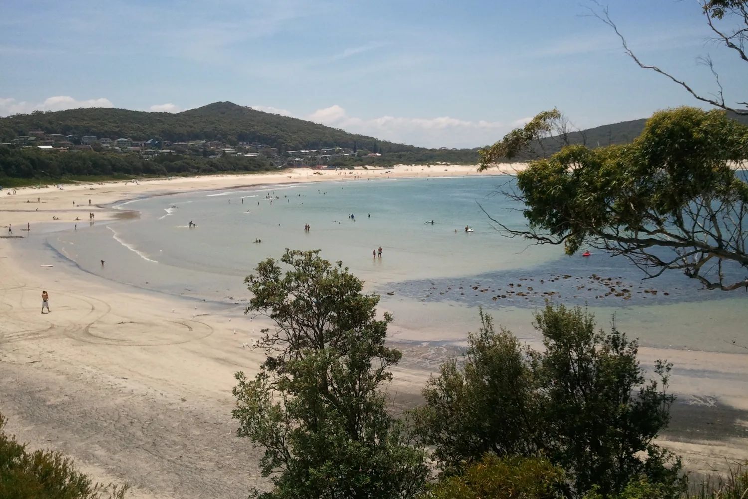 Fingal Bay beach from above: a calm curve of pale sand and shallow water, with wooded hills behind