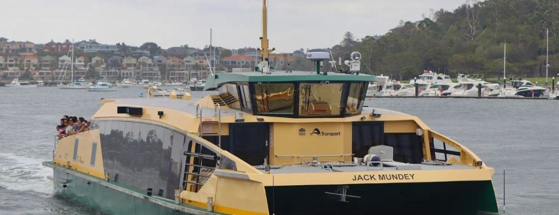 A green-and-gold Sydney ferry on the harbour, with moored boats and the far shore behind