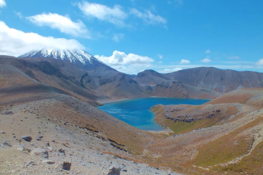 Upper Tama Lake in its explosion crater, blue water below red-brown slopes, with the snow cone of Ngāuruhoe behind.