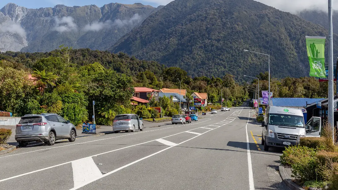 State Highway 6 running through Fox Glacier township, with parked cars, low buildings among trees and steep mountains behind.