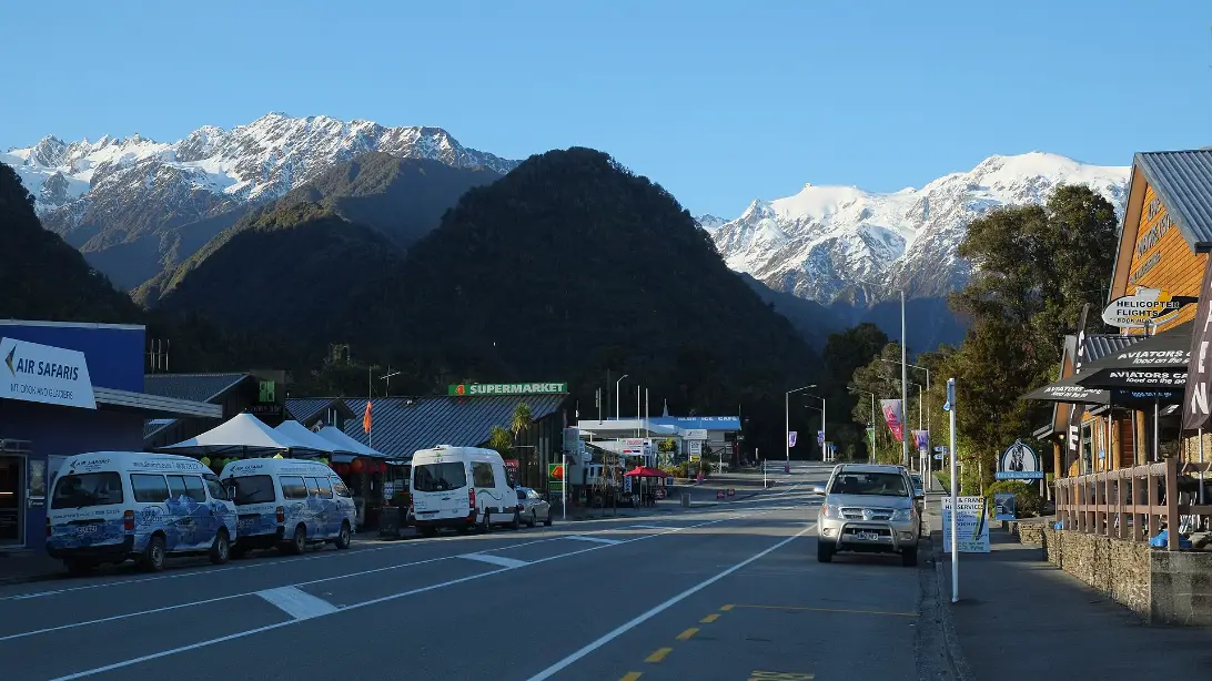 The main street of Franz Josef on a clear morning, with shops, parked tour vans and forested hills below snow-covered mountains.