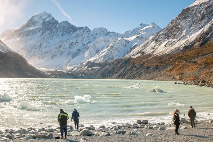 Walkers on the shore of Hooker Lake, with icebergs in grey water and the snow face of Aoraki / Mount Cook behind.