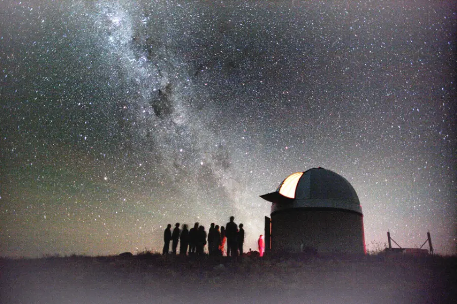 A group of people silhouetted beside an observatory dome under a sky full of stars and the Milky Way.