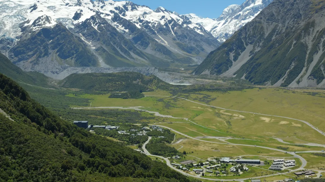 Mount Cook Village from the hillside above, a small cluster of buildings on the valley floor beneath steep slopes and snowy peaks.