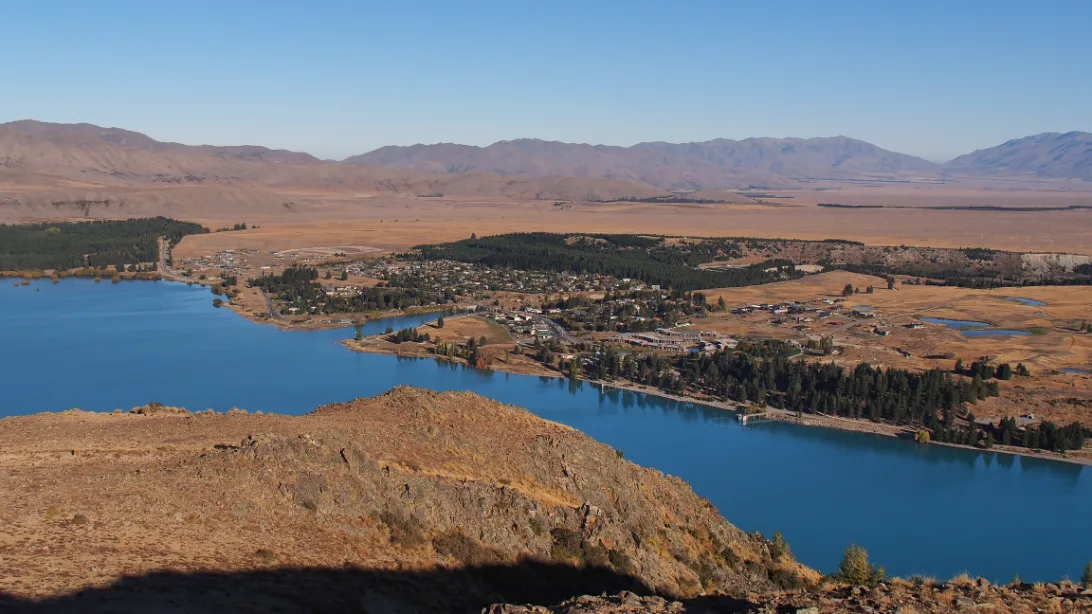 Lake Tekapo village seen from the hillside above, its houses and trees along the shore of the turquoise lake, with brown hills beyond.
