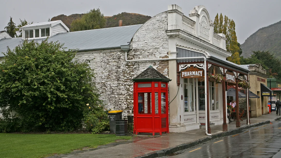 A whitewashed stone building with an ornate Victorian shopfront, now a pharmacy, beside a red telephone box on a wet street in Arrowtown, with café tables under the veranda beyond.