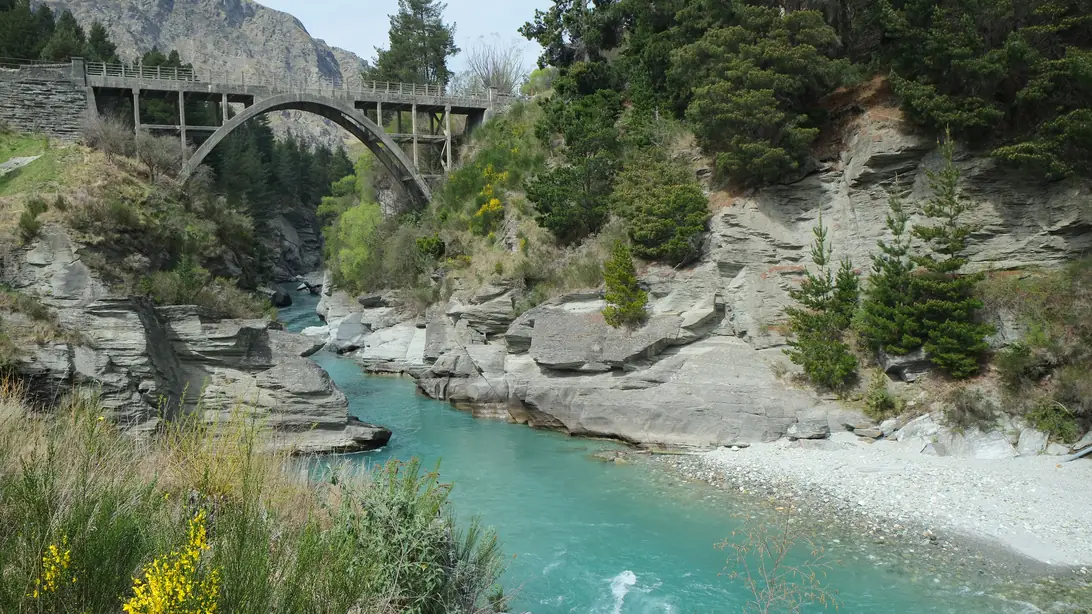 The turquoise Shotover River running through a rocky gorge at Arthurs Point, with the arch of the Edith Cavell Bridge above it.