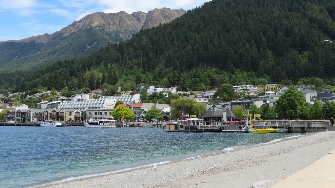 The small beach and waterfront of central Queenstown, with low buildings along the shore and a steep forested hill behind.