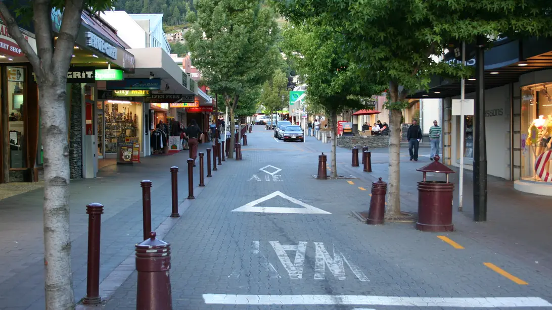 A street in central Queenstown lined with shops under trees in leaf, with bollards along the kerb and a forested hillside beyond.