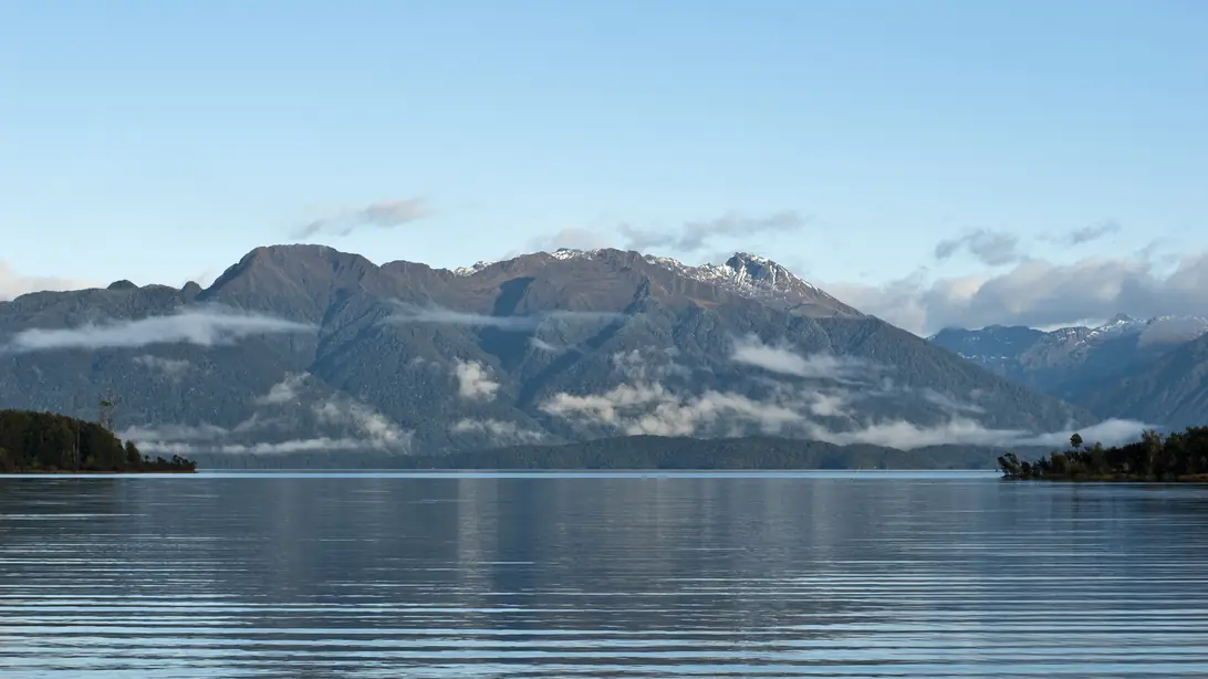 Lake Te Anau at Te Anau Downs, flat calm, with low cloud lying across forested mountains on the far shore.