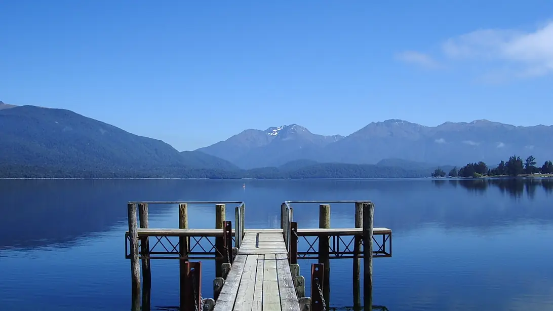 A wooden jetty on the Te Anau lakefront with the calm lake and the Fiordland mountains beyond, under a clear blue sky.