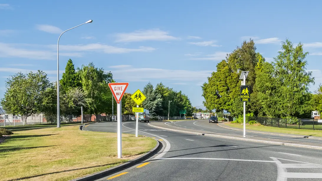 A road junction in the centre of Te Anau with a give-way sign, trees in leaf and a wide, quiet road under a blue sky.